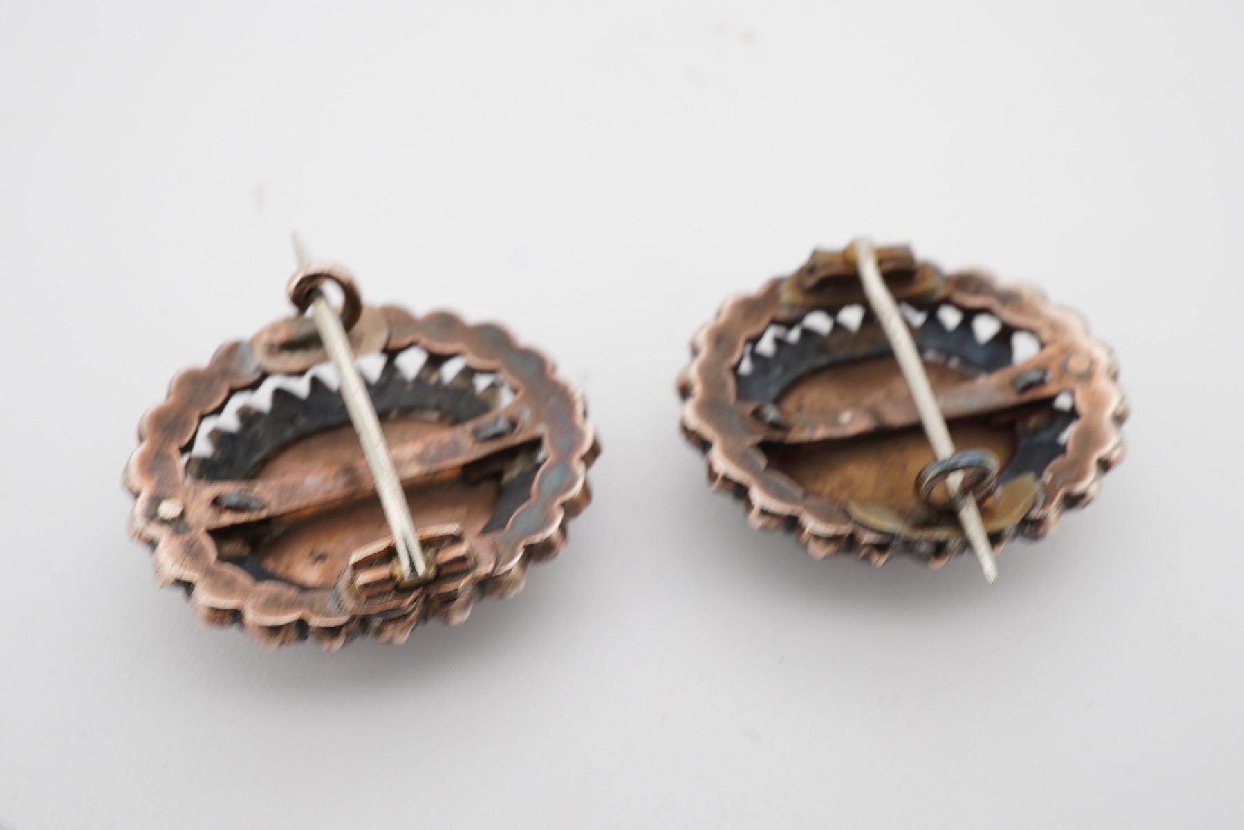A pair of early 20th century yellow metal mounted brooches, with central oval panel painted with the bust of a lady and bordered by concentric rows of seed pearls and garnets (adapted), 24mm by 20mm.
